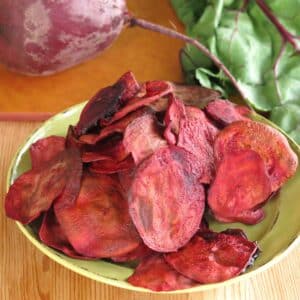 Baked beet chips piled on a green plate with a raw beet with stem in place behind it.