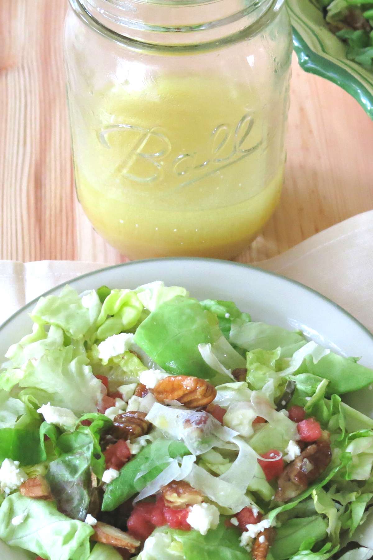 Orange honey mustard vinaigrette in a mason jar with a plate of summer salad with fennel, watermelon, feta, and candied pecans in front of it.