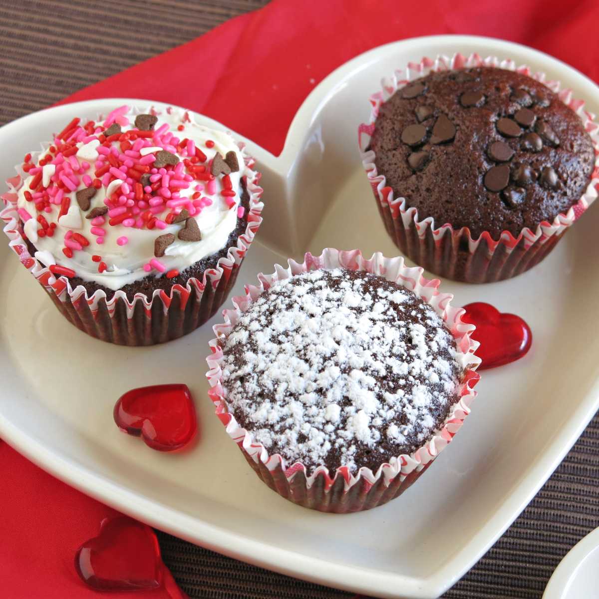Three decorated eggless chocolate cupcakes: one with chocolate chips on top, one with powedered sugar, and one with vanilla frosting and sprinkles.