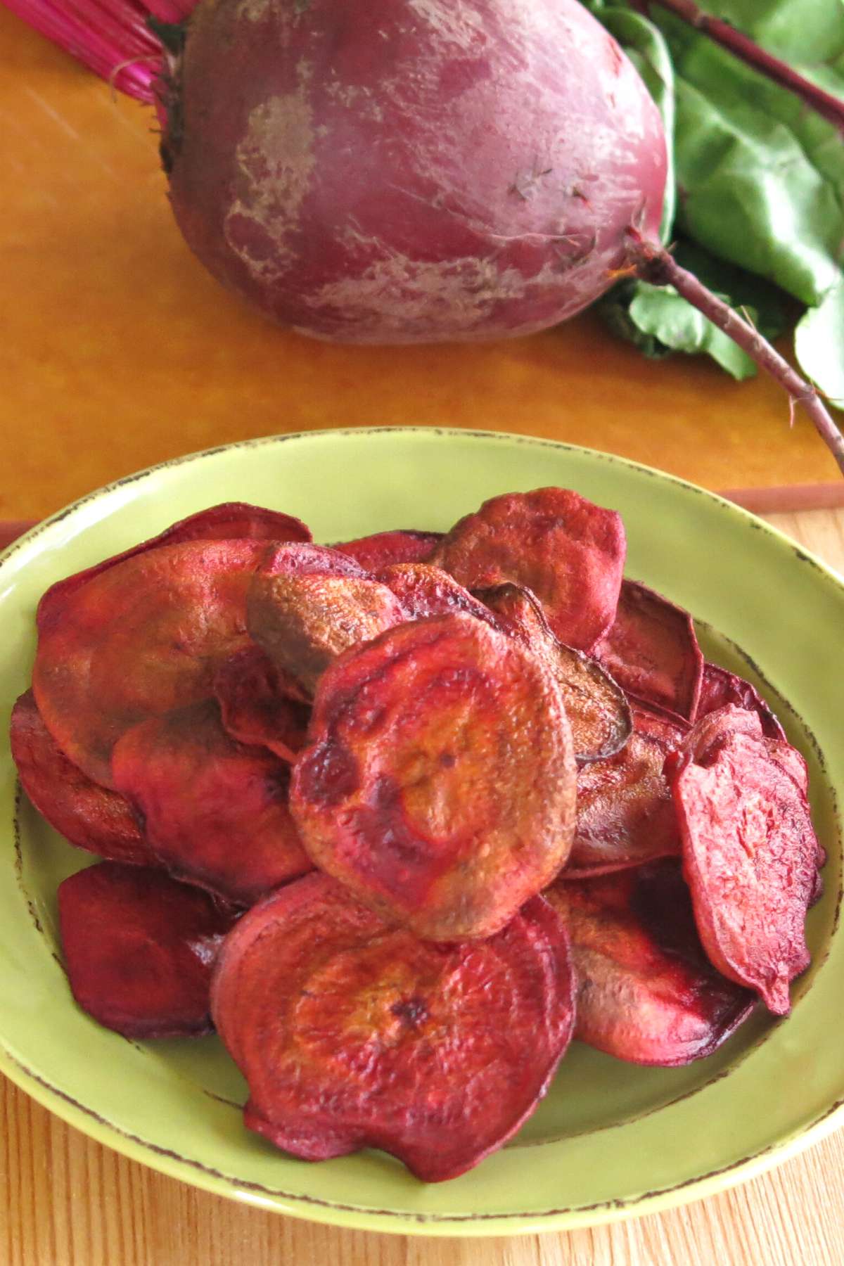 Homemade baked beet chips piled on a plate with a raw beet behind them on a cutting board.