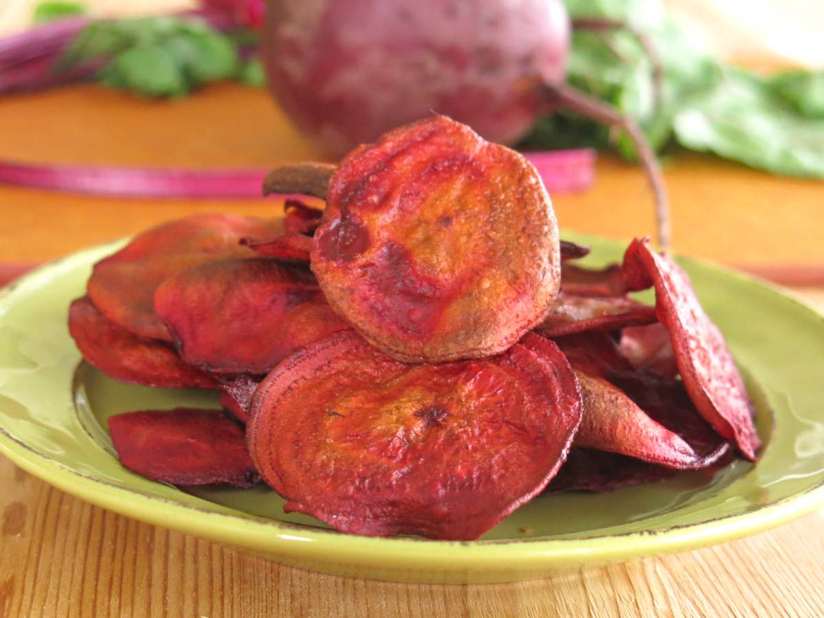 Oven-baked beet chips stacked on a plate with a raw red beet with the stem on behind it.
