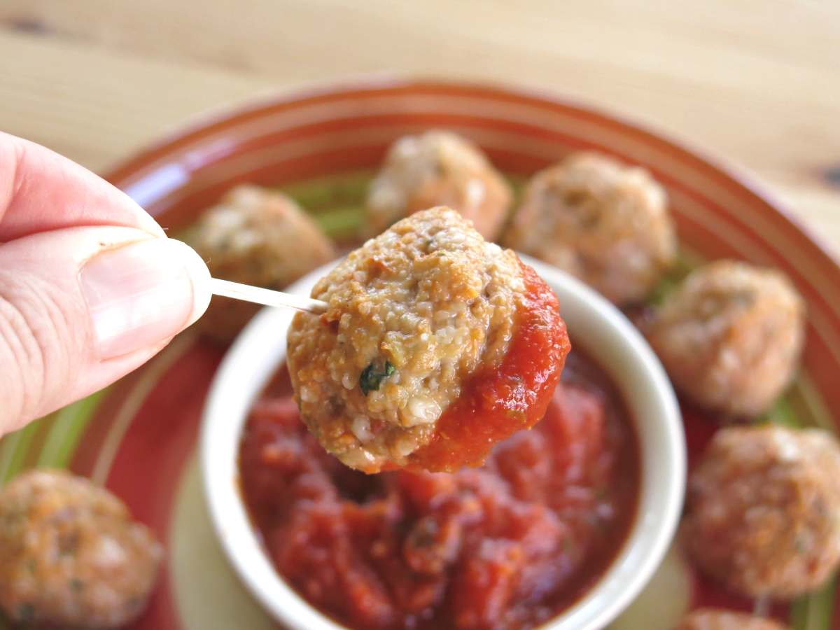 Fingers holding a parmesan chicken meatball on a toothpick over a bowl of marinara surrounded by more meatballs.