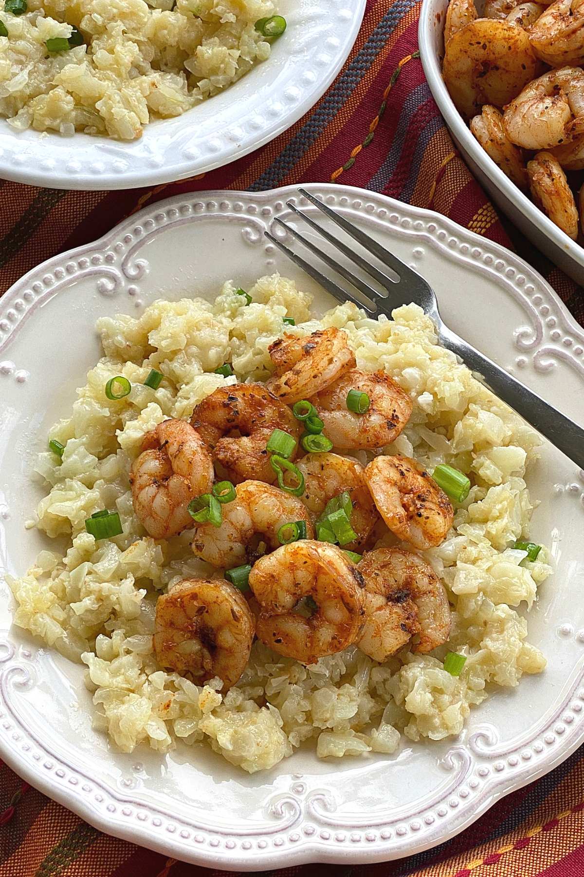 Blackened shrimp on top of a plate with cauiflower grits with more shrimp  and a bowl of grits in the background.