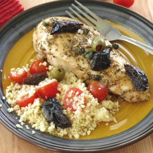 A plate with chicken marbella made with boneless chicken breasts that is topped with prunes, olives, tomatoes, with a side of couscous next to it.