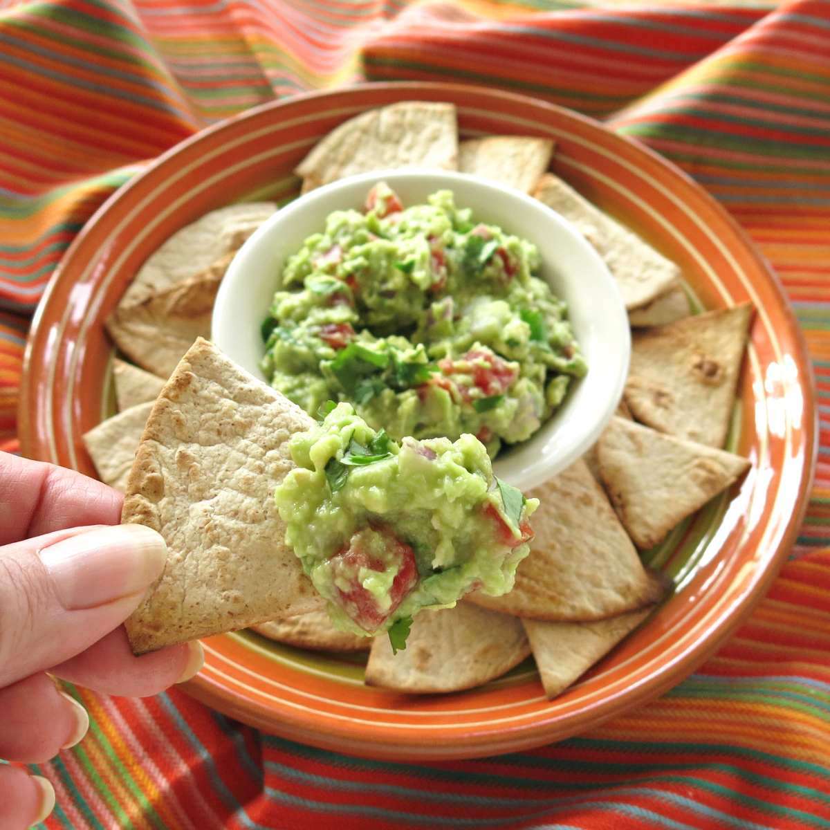 Low-carb tortilla chips on a plate surrounding a bowl of guacamole with fingers holding up a chip with some dip on it.