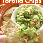 Low-carb tortilla chips on a plate around guacamole with fingers holding one with dip on it.