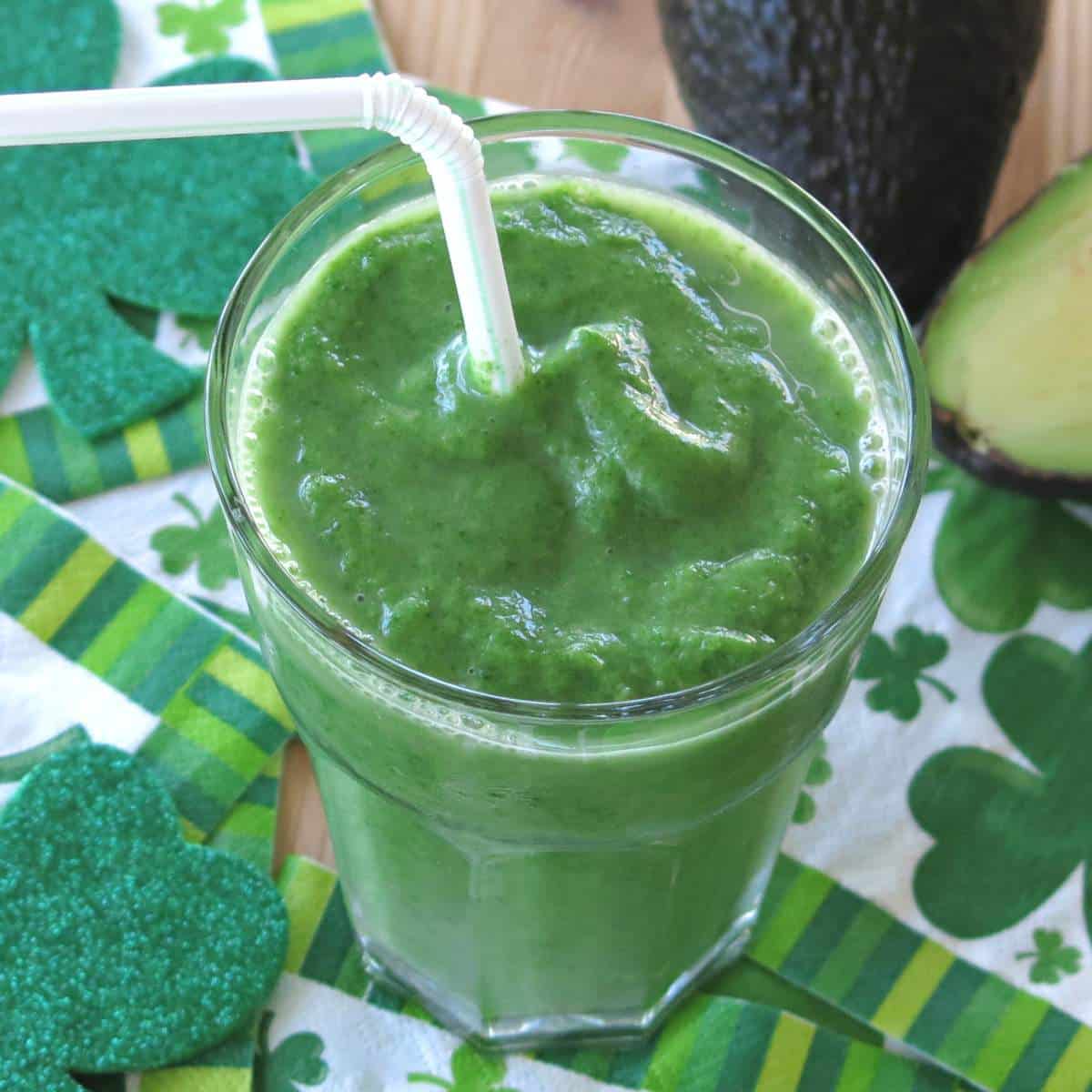 Pineapple avocado smoothie with spinach and ginger in a glass with a straw with an avocado behind it.