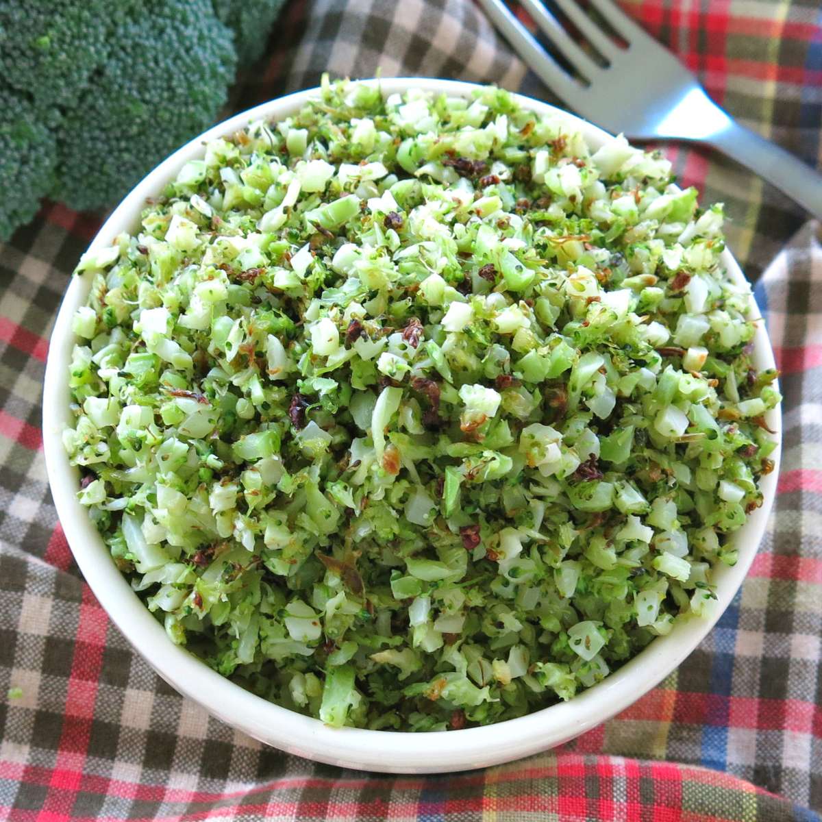 Riced broccoli that has been roasted in bowl next to a head of broccoli and a fork.