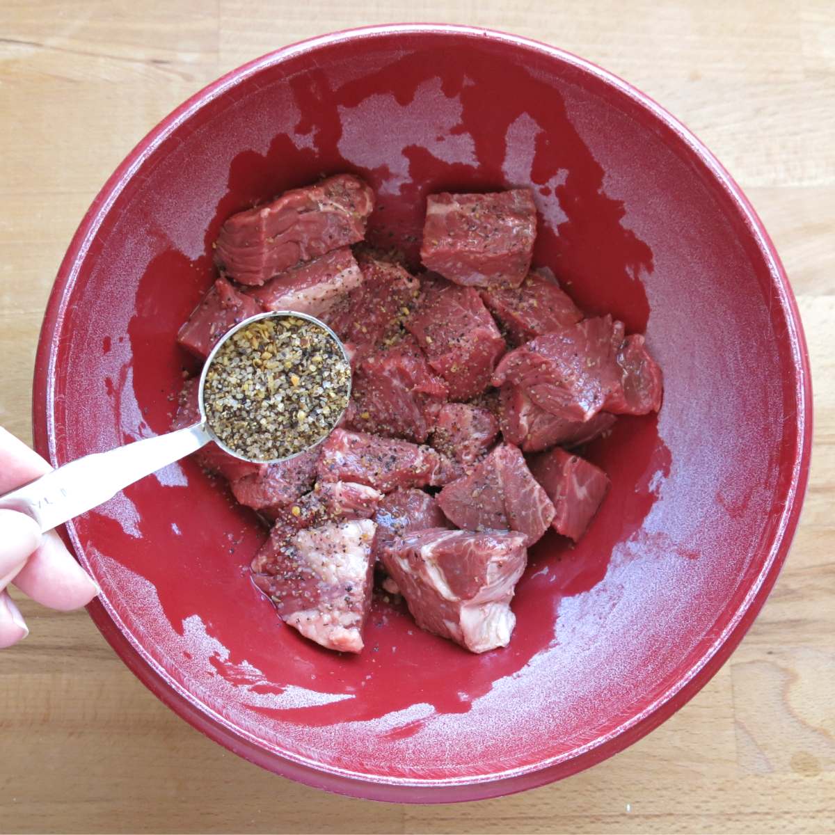Seasoning in a measuring spoon being sprinkled over cubed beef in a red bowl.
