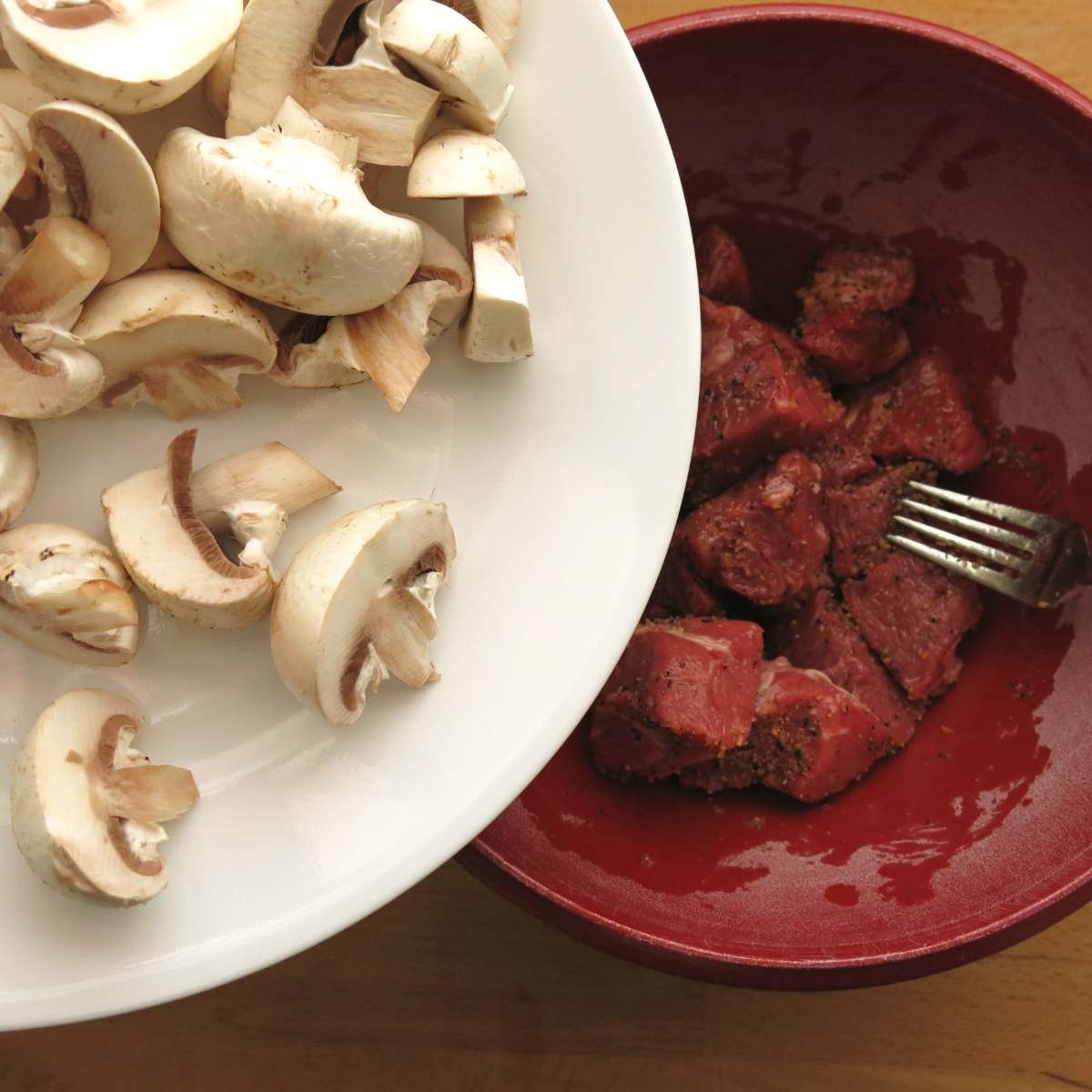 Chopped mushrooms on plate being added to bowl with seasoned beef cubes.