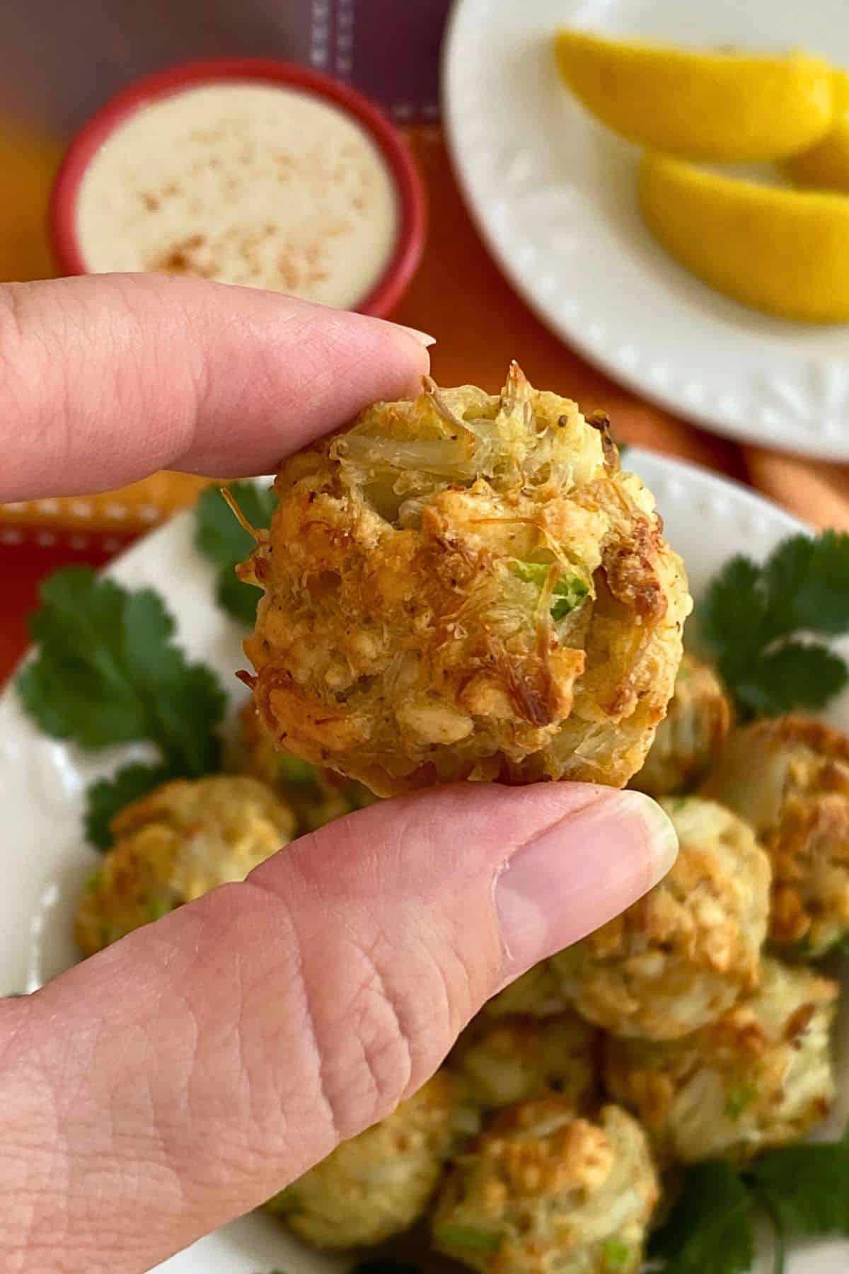 Fingers holding a crab ball appetizer over a bowl with more.