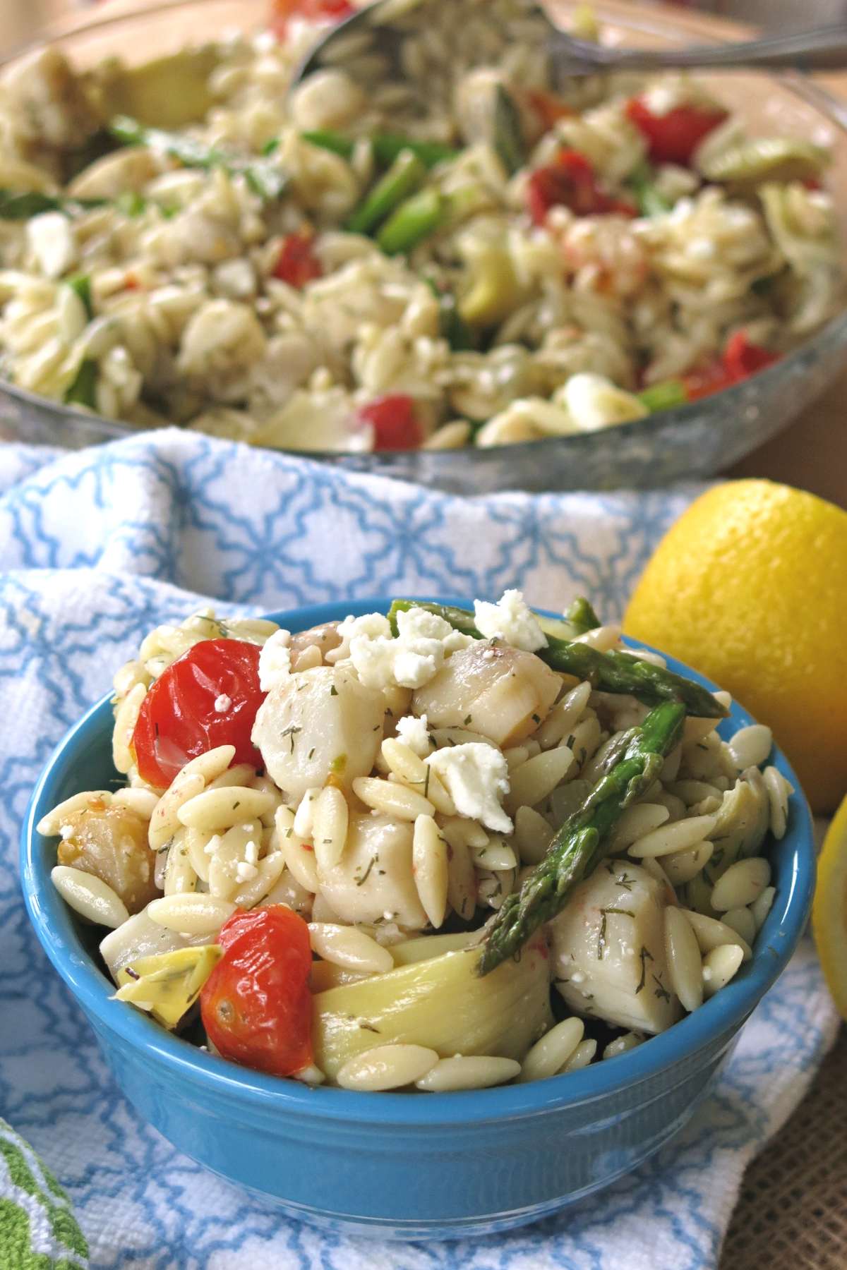 Mediterranean warm orzo salad with scallops, asparagus, artichokes, tomatoes, and feta cheese in a bowl. with serving dish behind it.