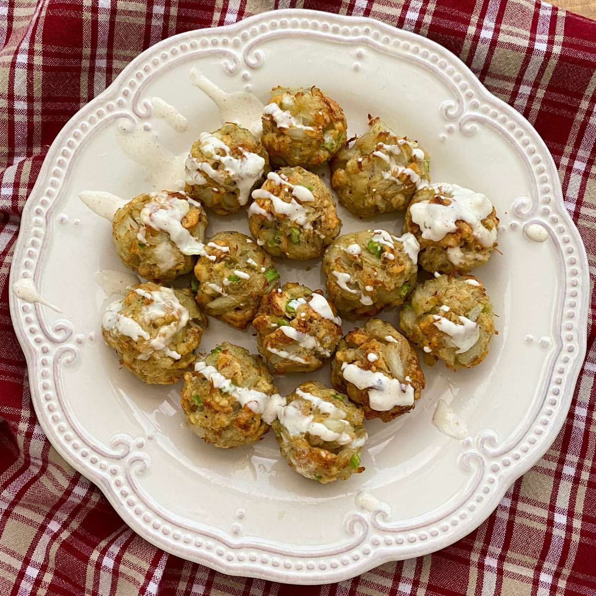 Mini crab cake bites on a plate drizzle with lemon garlic aioli and a bowl with more crab balls and aioli behind them.