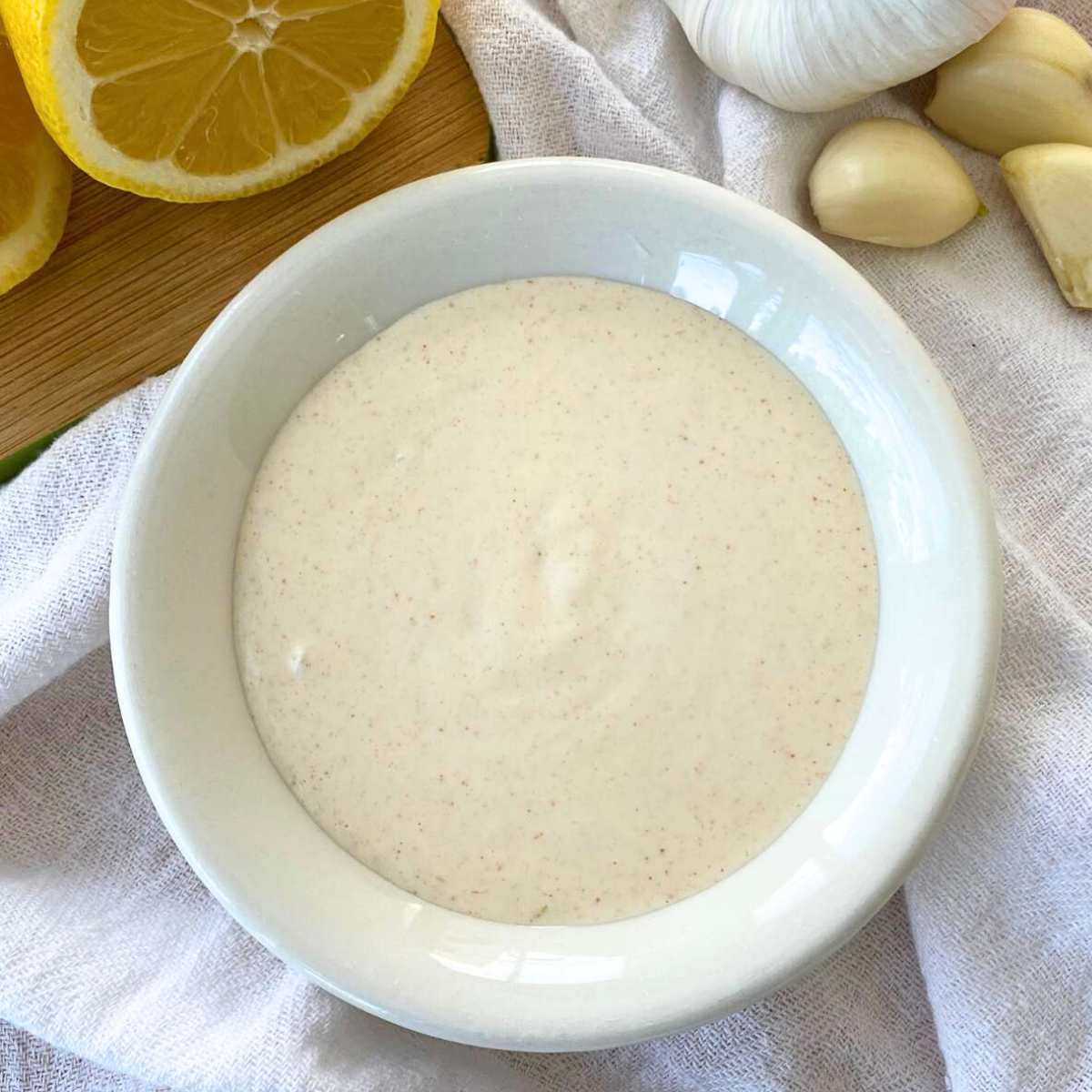 Lemon garlic aioli sauce in a bowl with a cut lemon and garlic cloves behind it.