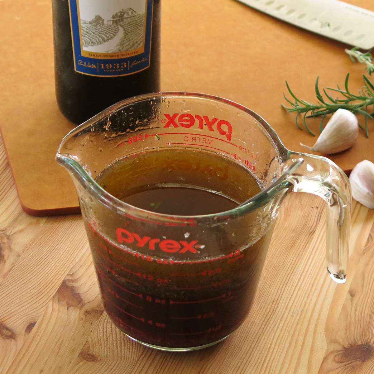 Red wine steak marinade in a glass measuring cup with a bottle of red win, garlic cloves, and fresh rosemary next to it.