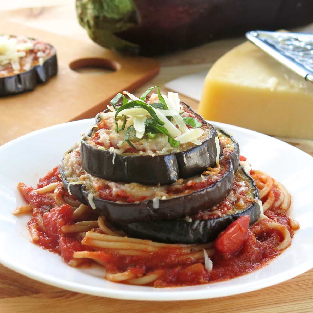 Eggplant Parmesan Stack on a white plate with an eggplant and triangle of Parmesan cheese behind it.