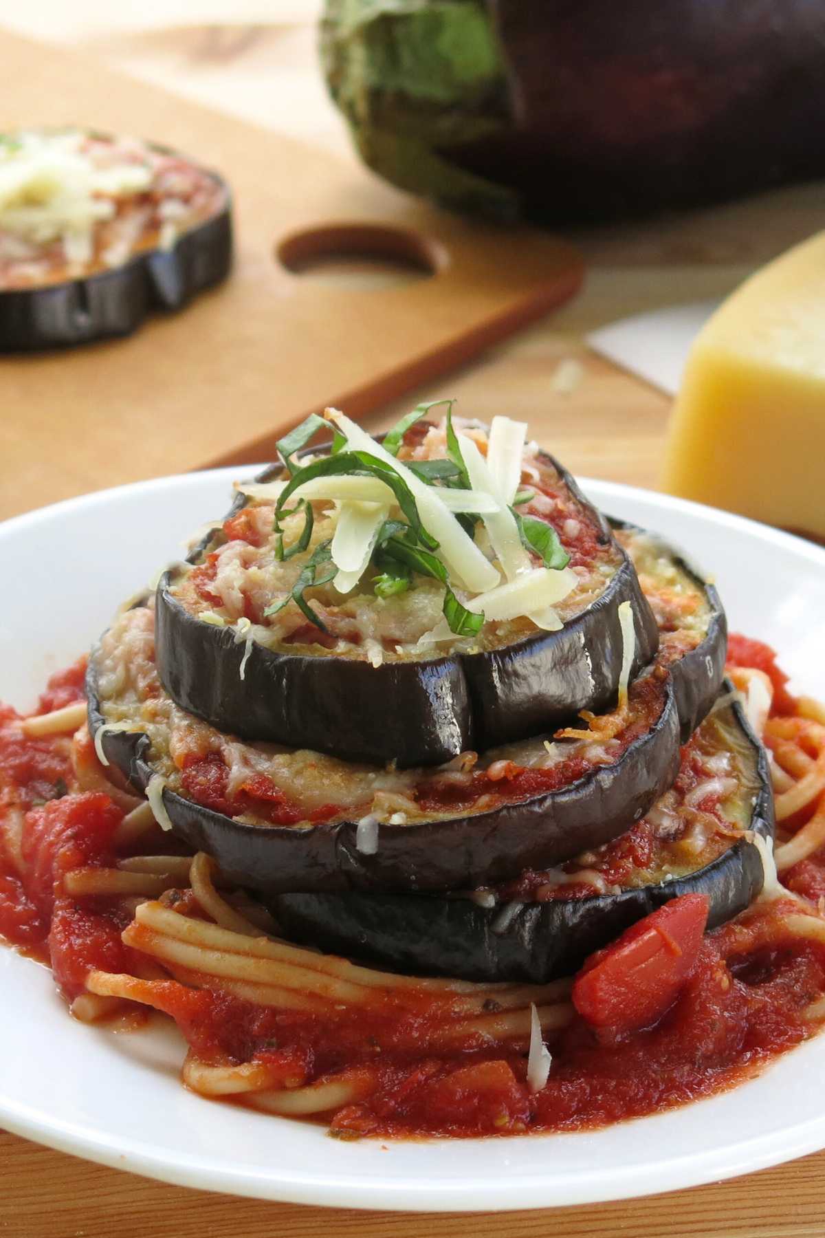 Gluten-free eggplant parmesan stacks on a plate with an eggplant and a cooked eggplant slice behind it.