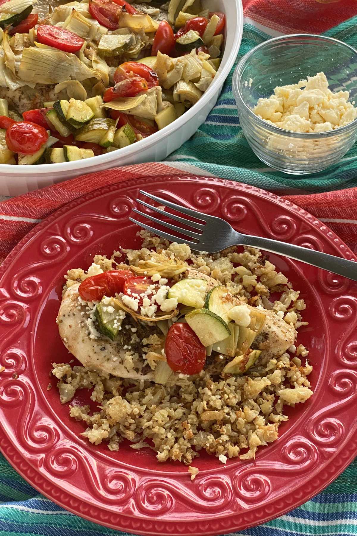 Mediterranean baked chicken topped with feta cheese on a red plate with platter of the dish and cup of feta cheese behind it.