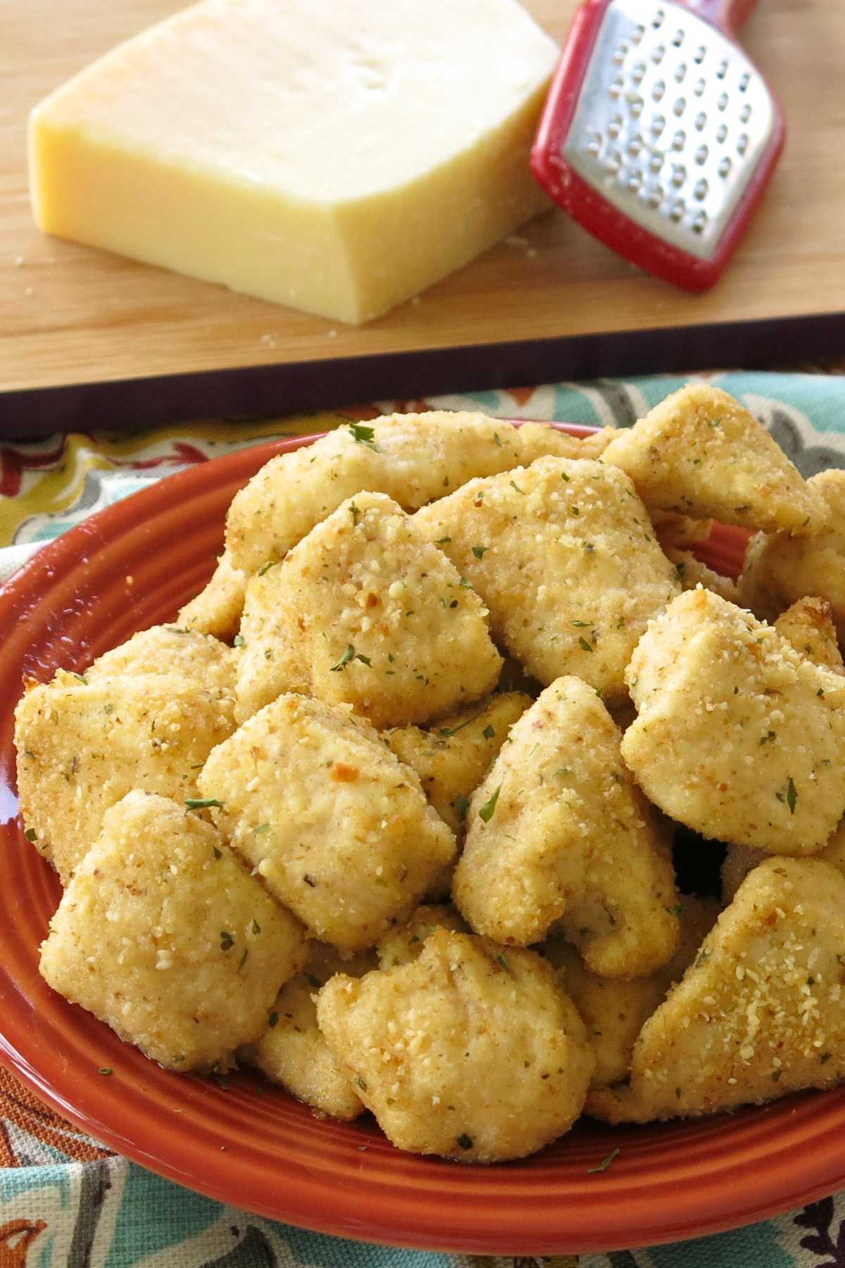 Garlic parmesan chicken bites piled on a plate with a block of parmesan cheese and grater behind it.