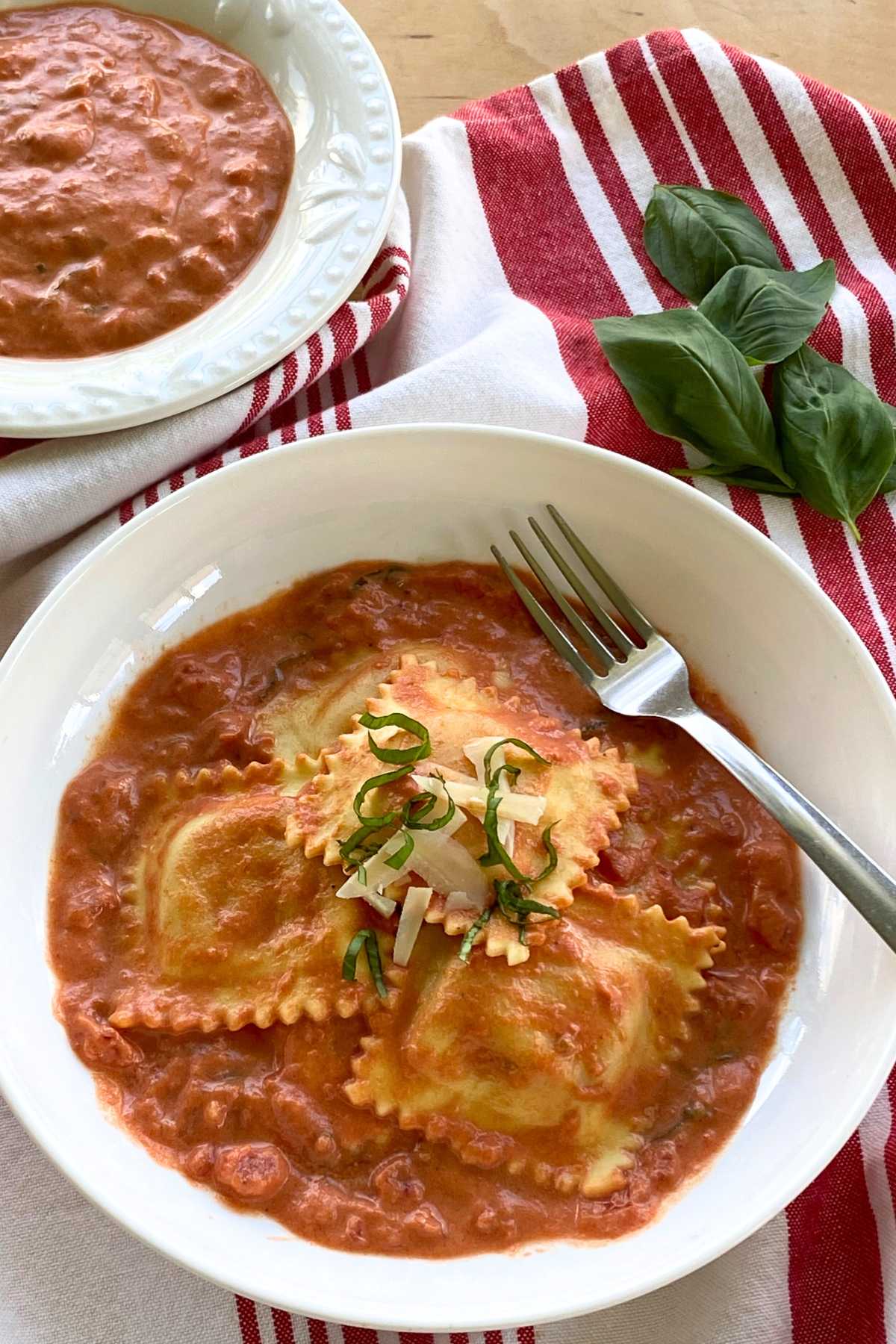 Creamy pink lobster ravioli sauce in bowl with lobster ravioli with a bowl of sauce and fresh basil leaves behind it.