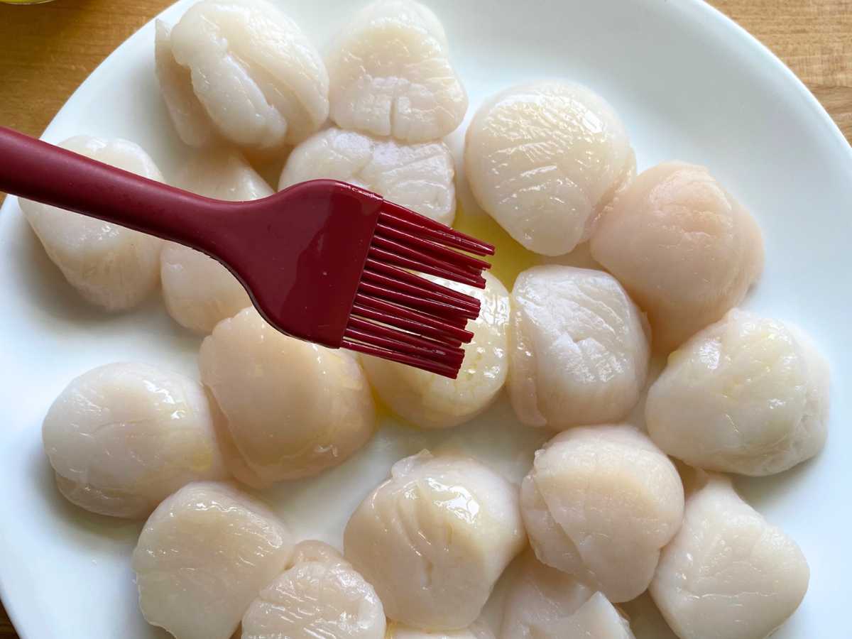 Silicone brush applying butter and olive oil mixture to sea scallops on a plate.