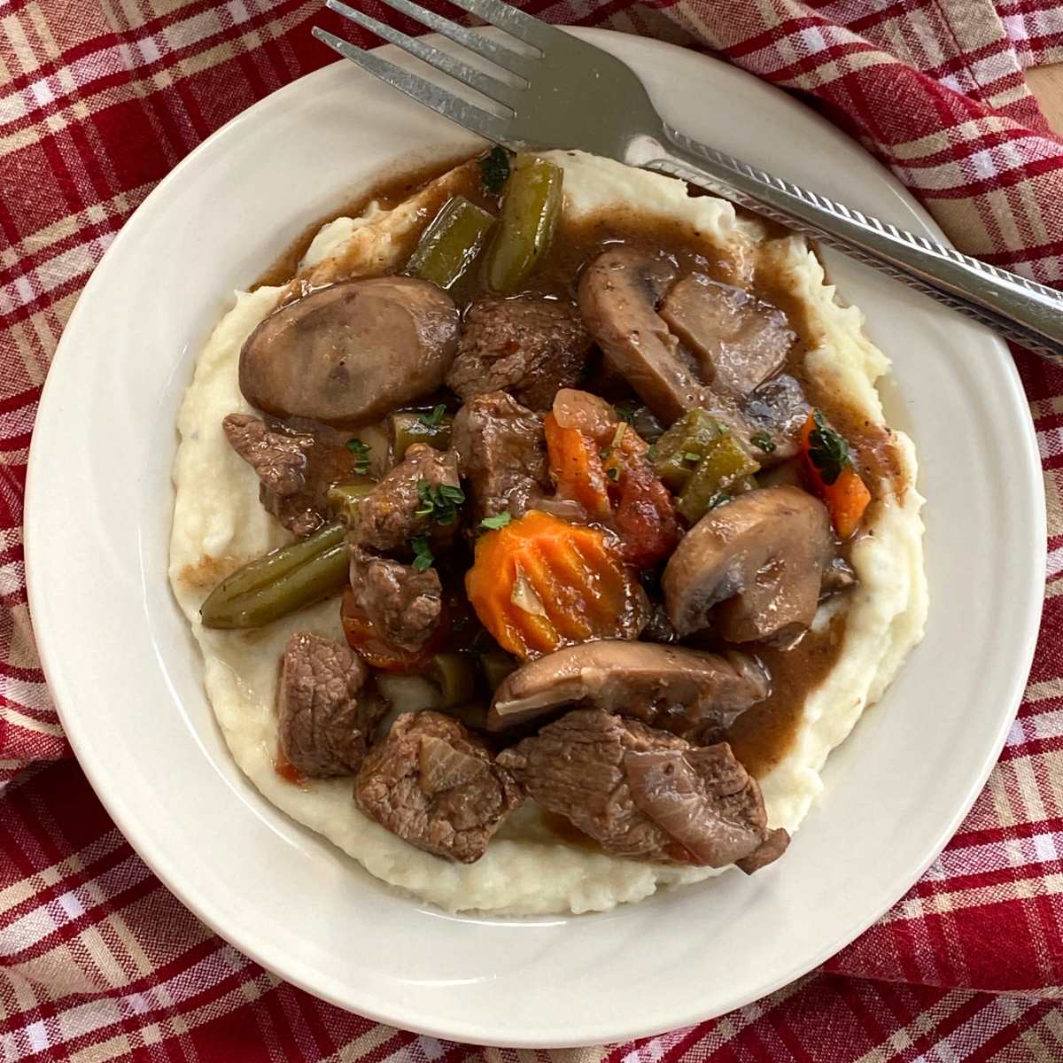 Crock-pot beef stew with mushrooms, carrots, onions, and green beans on top of mashed potatoes on a plate with lots of red wine sauce.