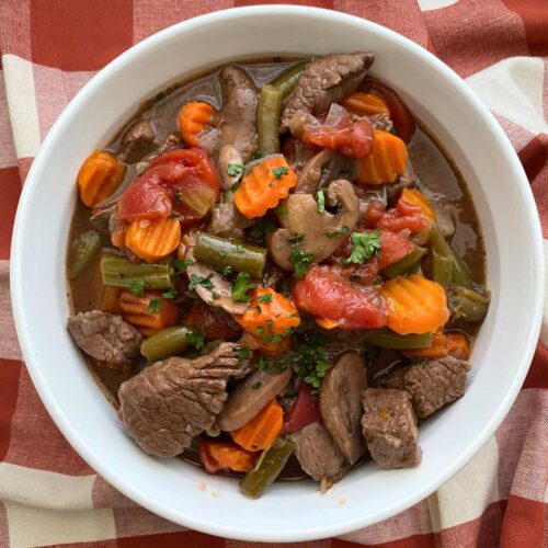 Large bowl of crock-pot beef stew in red wine sauce with mushrooms, tomatoes, carrots, and green beans.