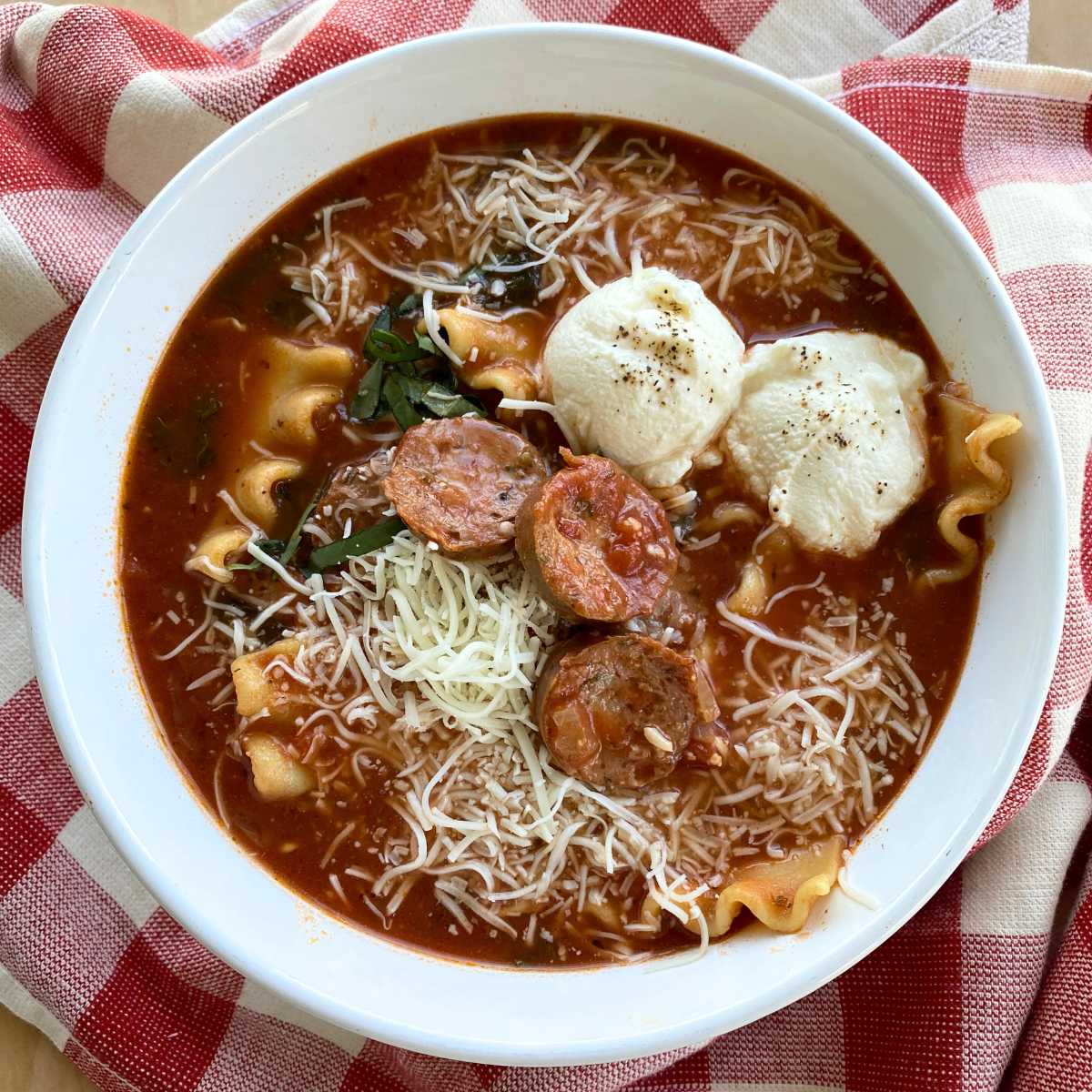 One pot lasagna soup topped with two dollops of ricotta cheese and garnished basil and more mozzarella cheese in a white bowl on a red checkered napkin.