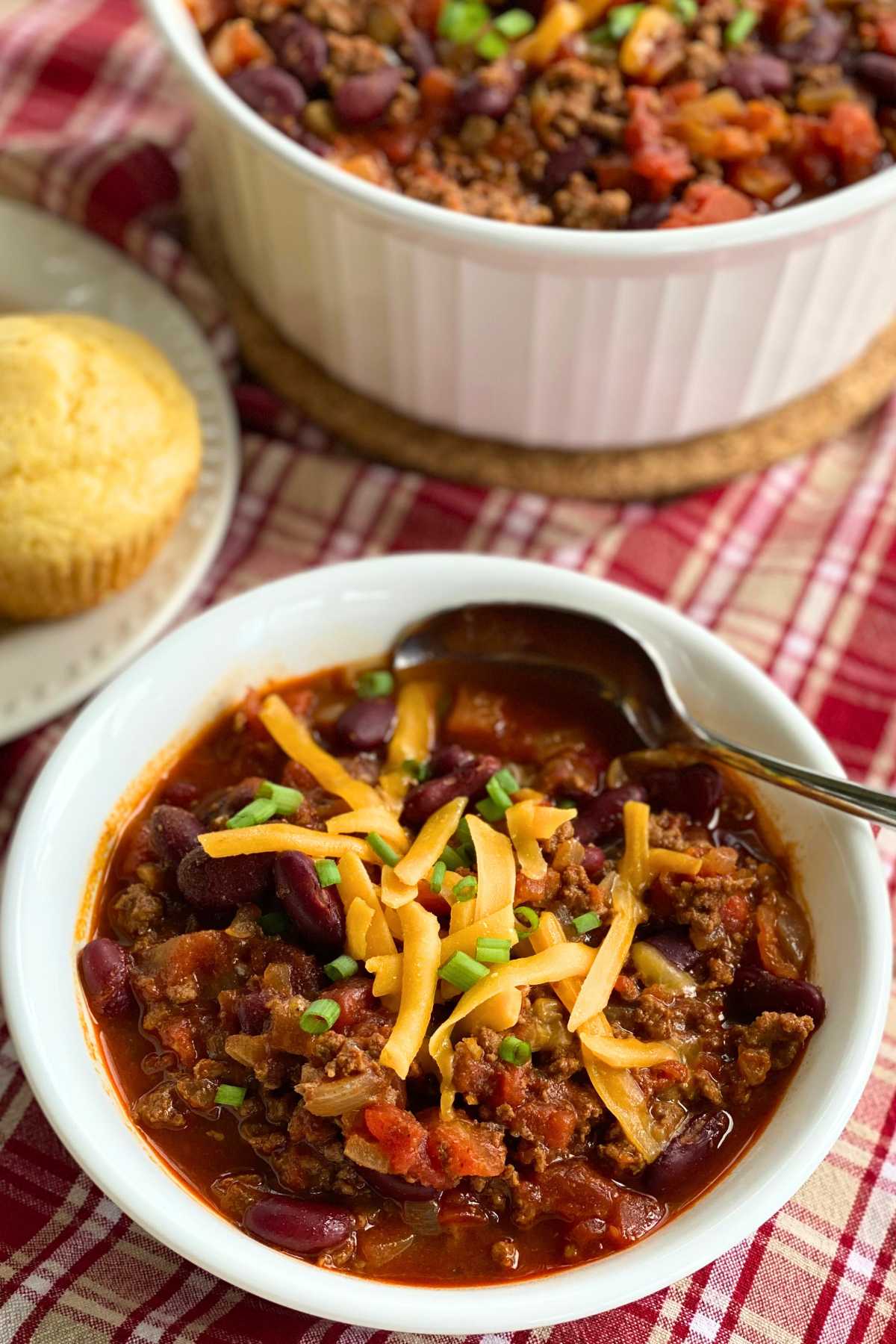 Individual bowl of easy chili with beef and beans topped with shredded cheddar cheese and green onion with cornbread muffins behind it.