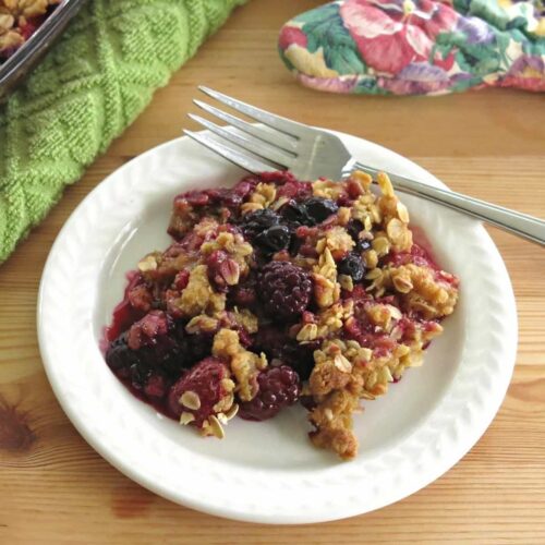 Triple berry crisp with blueberries, strawberries, and black berries on a plate with a fork. The oatmeal crumb topping is browned.