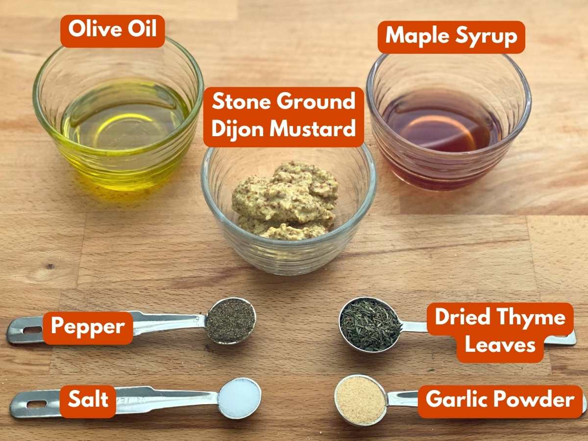 Olive oil, stone ground mustard, maple syrup, pepper, salt, dried thyme leaves, and garlic powder in glass cups or measuring spoons with a text overlay with the name of each ingredient.