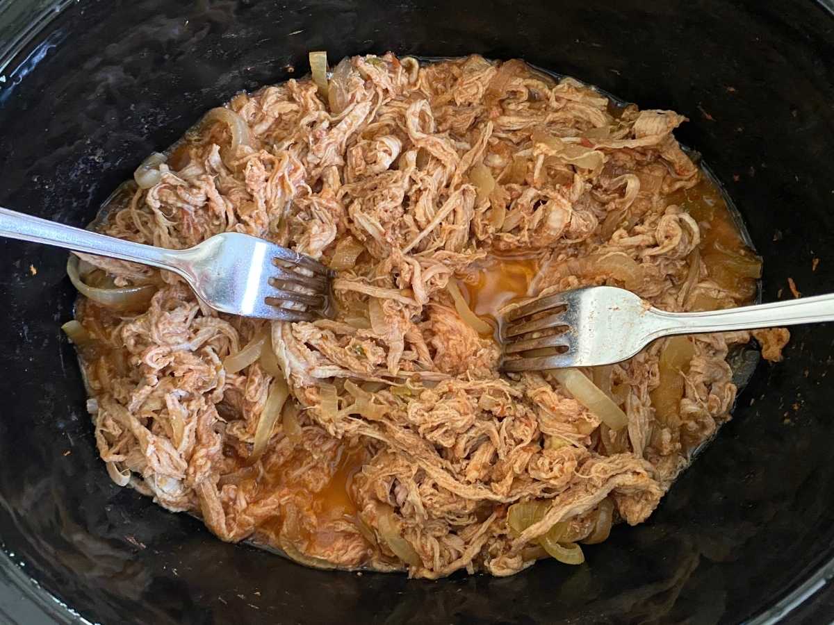 Cooked pork carnitas in the base of slow cooker with two forks.