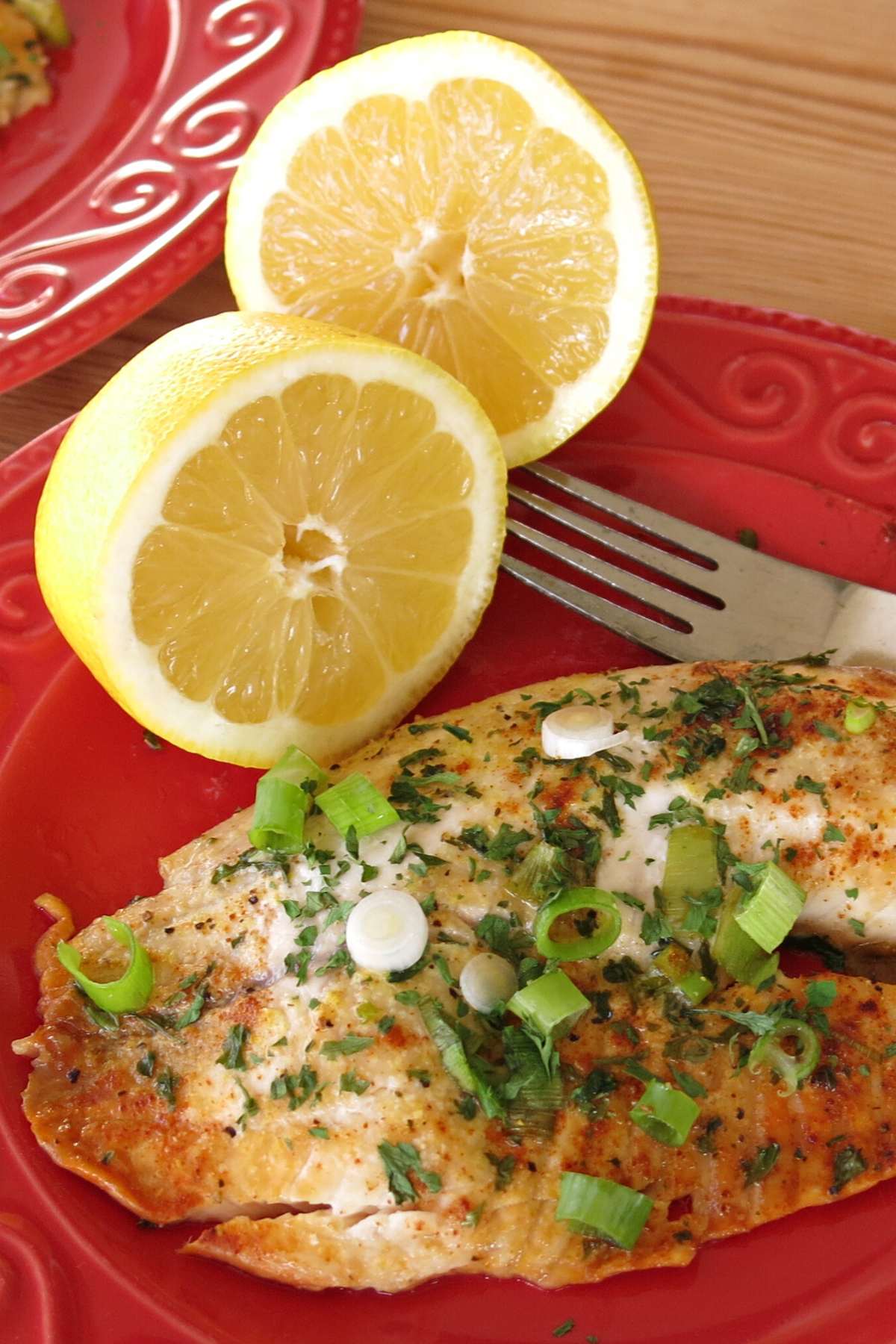 Oven-baked lemon pepper tilapia on a red plate with a fork and a lemon that has been cut in half.