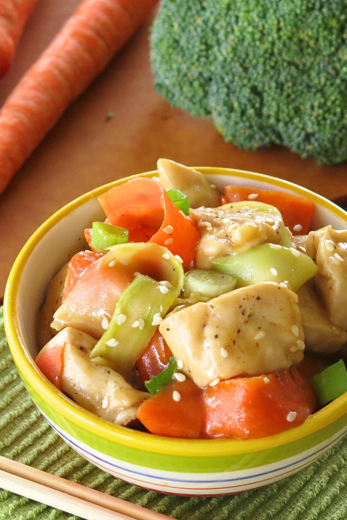 Healthy sweet and spicy orange chicken with shaved broccoli and carrots in bowl with a head of broccoli and carrots behind the bowl.