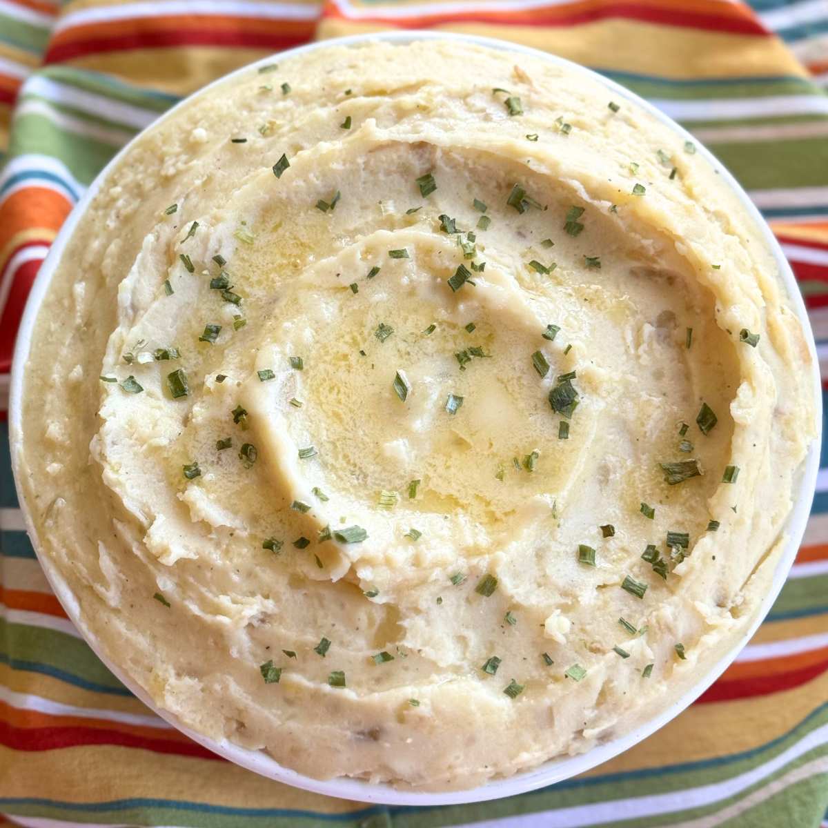 Creamy crock-pot mashed potatoes with cream cheese and sour cream in a serving bowl with pools of butter, and chives on top.