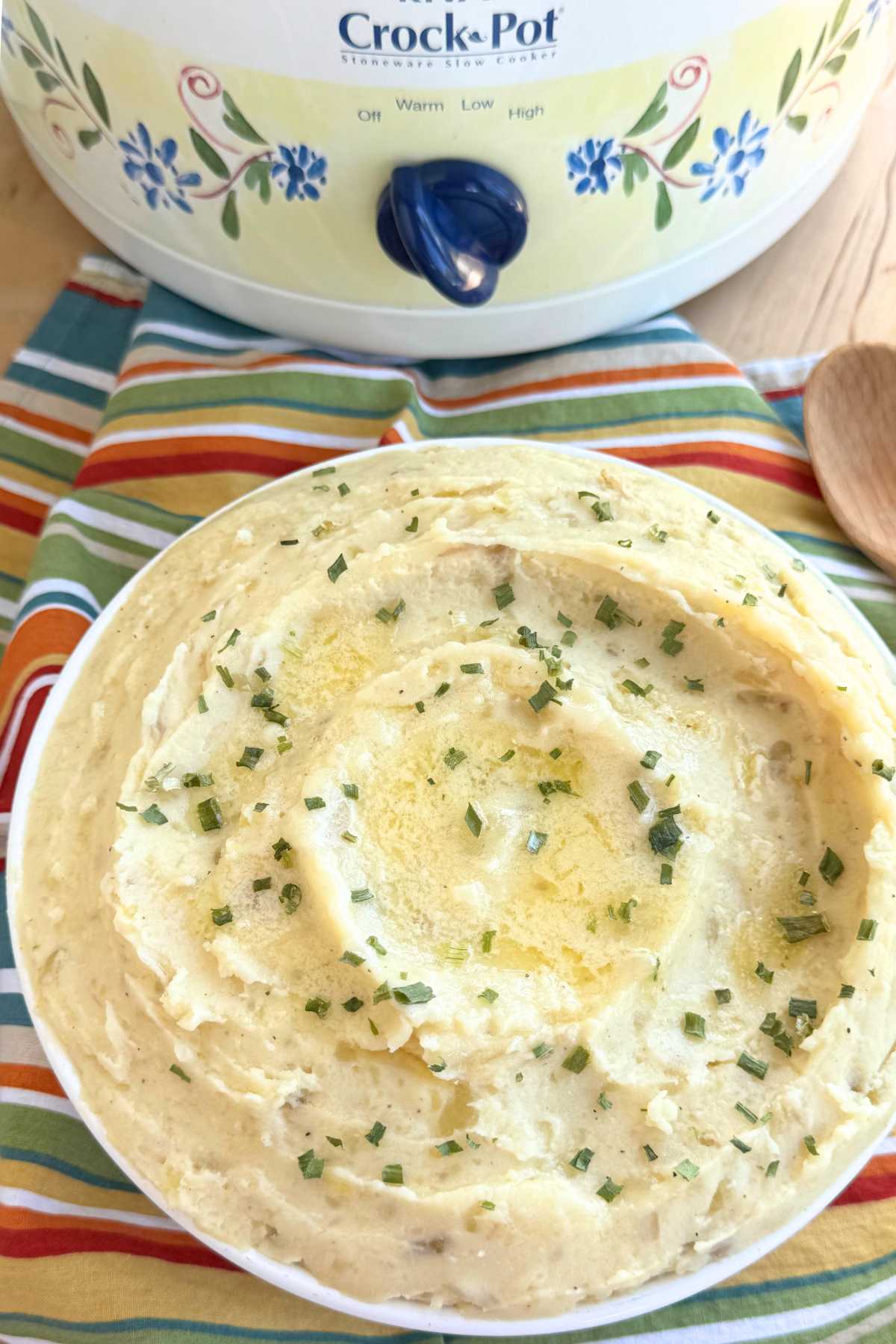 Creamy slow cooker mashed potatoes topped with melted butter and chives in a large bowl with a slow cooker behind it.