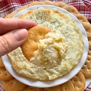 Easy artichoke dip with cream cheese on a cracker over a serving platter with more dip.