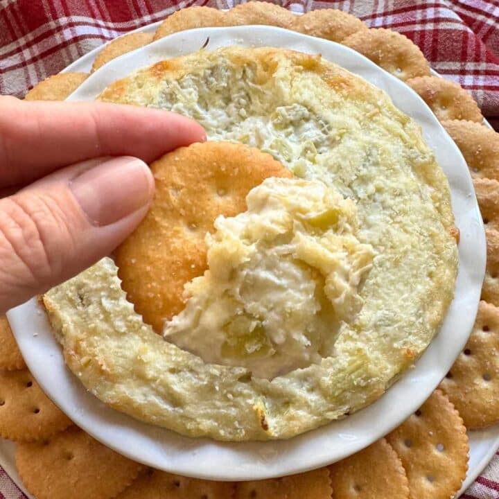 Easy artichoke dip with cream cheese on a cracker over a serving platter with more dip.