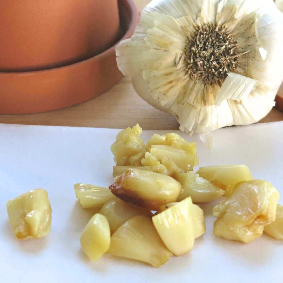 Roasted garlic cloves on parchment paper with a garlic head and garlic keeper behind them.