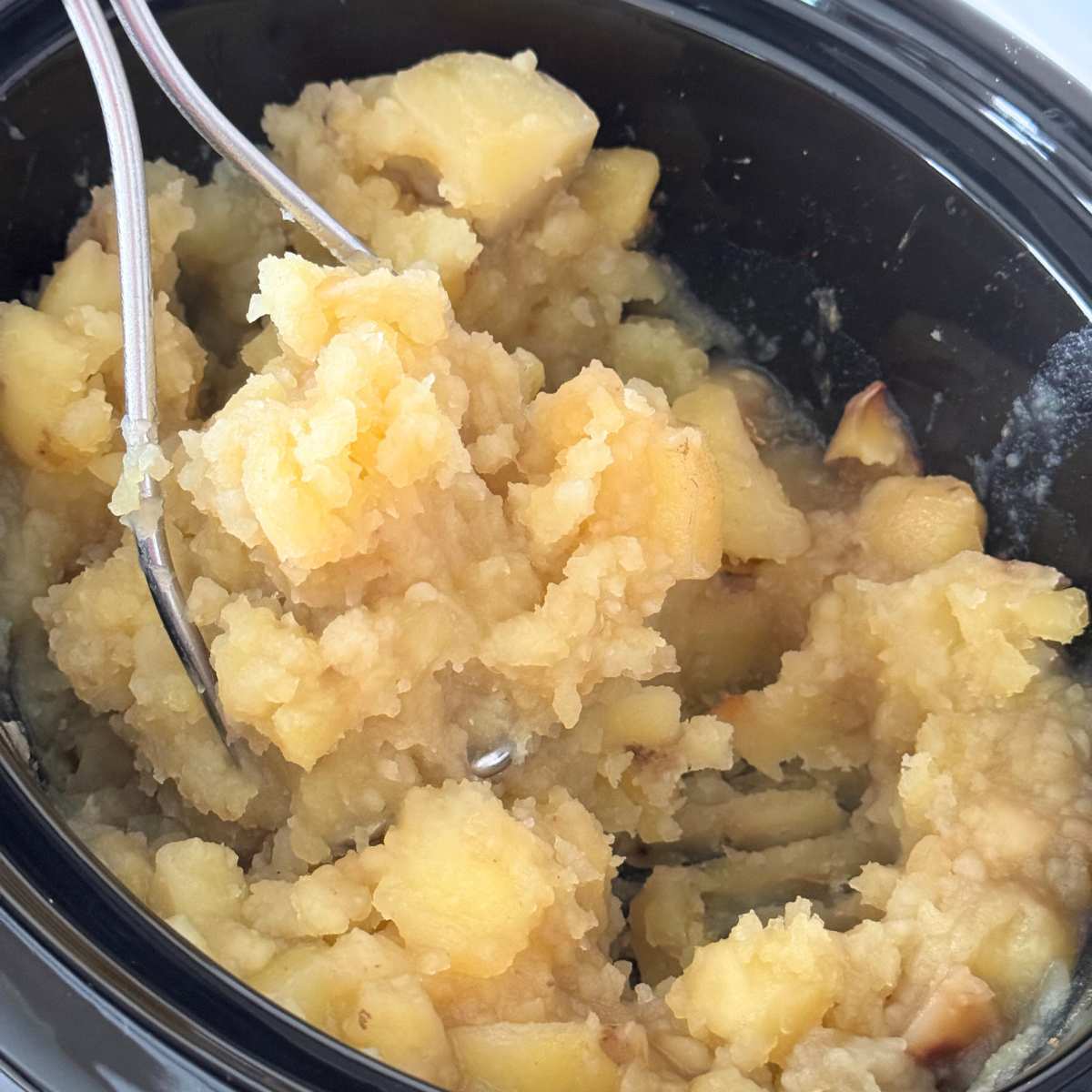 Cooked potatoes being mashed with a potato masher in the base of a crock-pot.