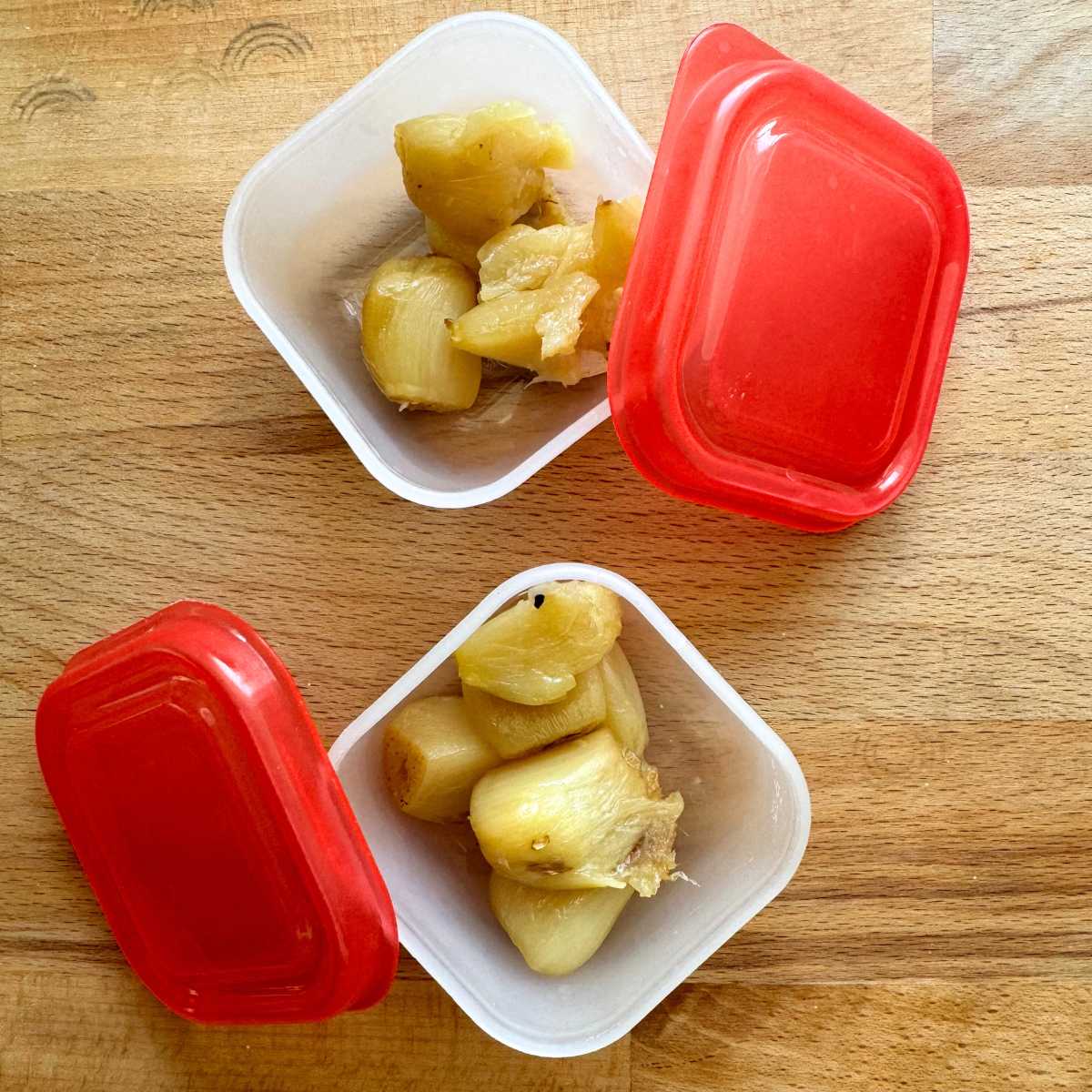 Two very small freezer containers with roasted garlic cloves inside. Red lids are leaning against the container.