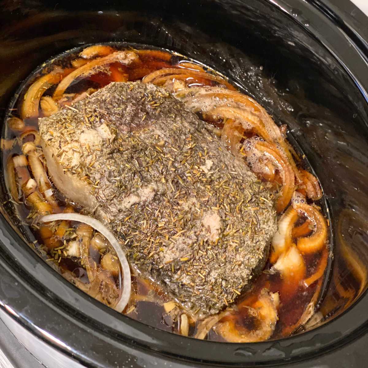 Cooked bottom round roast in a crock-pot surrounded by onions and beef broth.