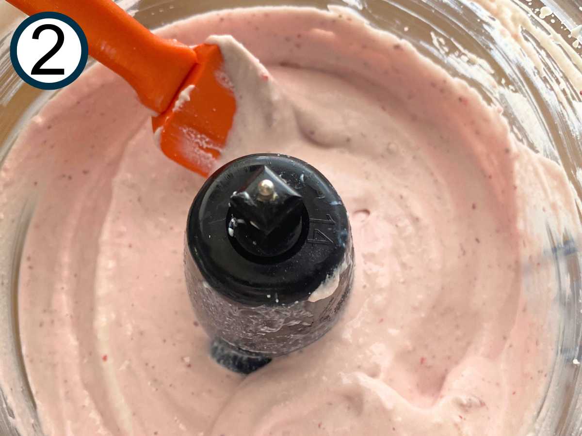 whipped cottage cheese, strawberries, vanilla extract, and sweeteners in the base of a food processor being stirred with a spatula.