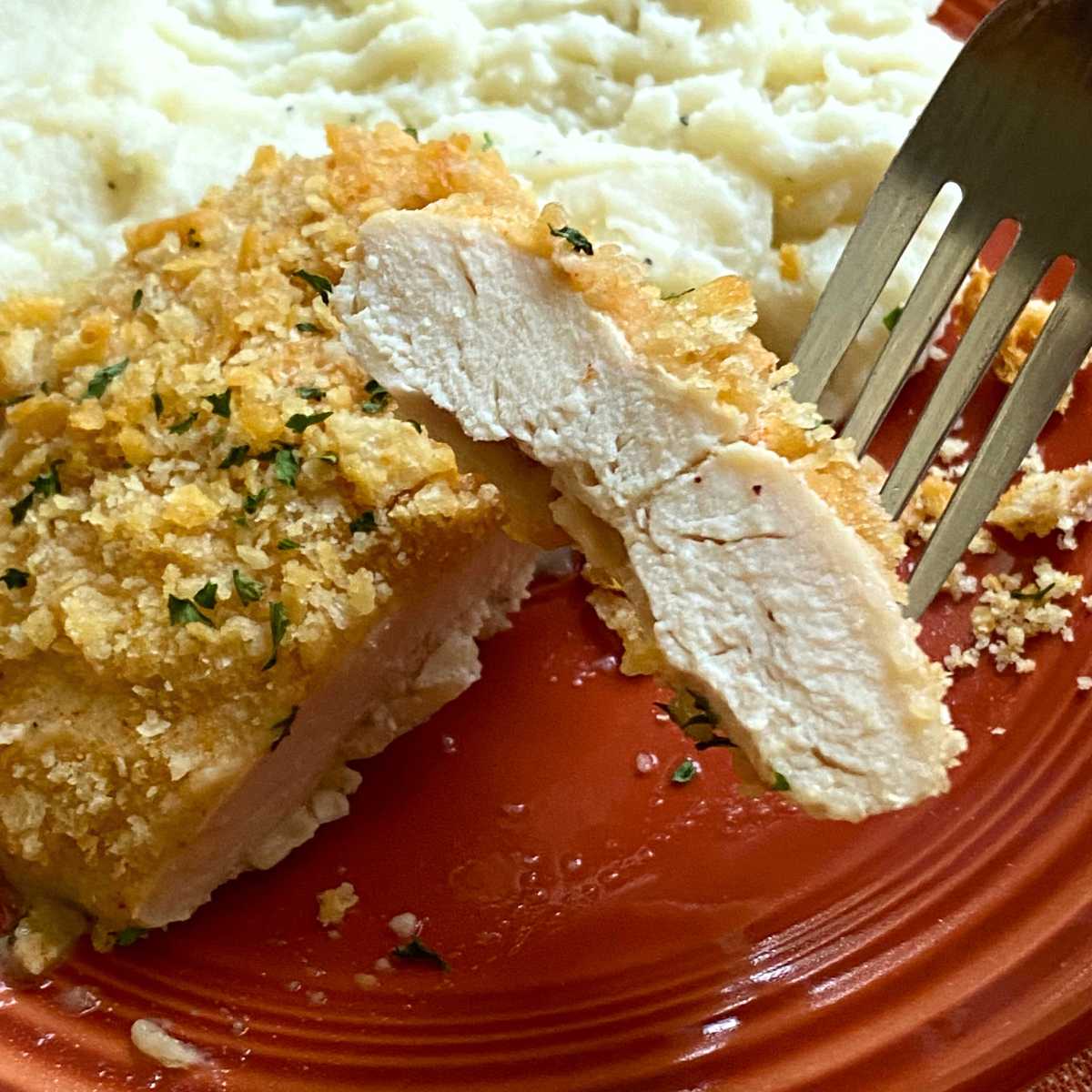 Fork holding a bite of heavenly chicken with the rest of the chicken breast coated with Ritz crackers on the plate with mashed cauliflower.