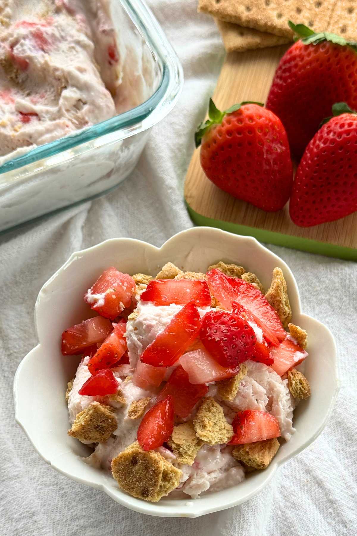 Strawberry cheesecake cottage cheese ice cream in a tulip bowl with fresh strawberries, graham crackers, and the container with more ice cream behind it.