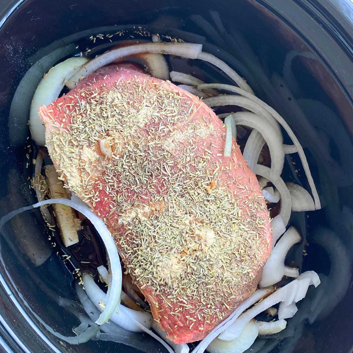 Uncooked bottom round roast seasoned with spices on top of onions and surrounded by beef broth in a crock-pot.