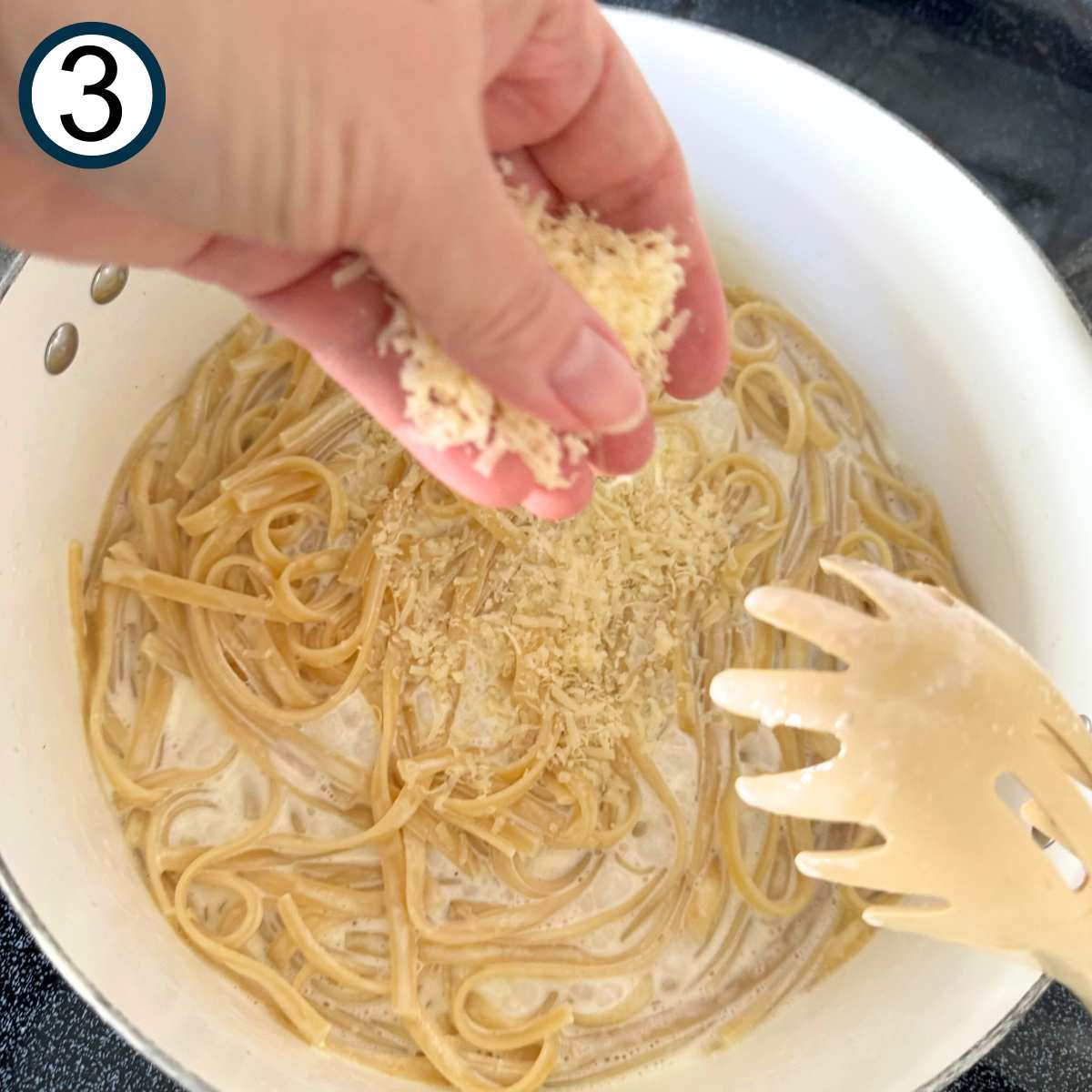 Hand dropping grated Parmesan cheese into a pot with pasta and cream.