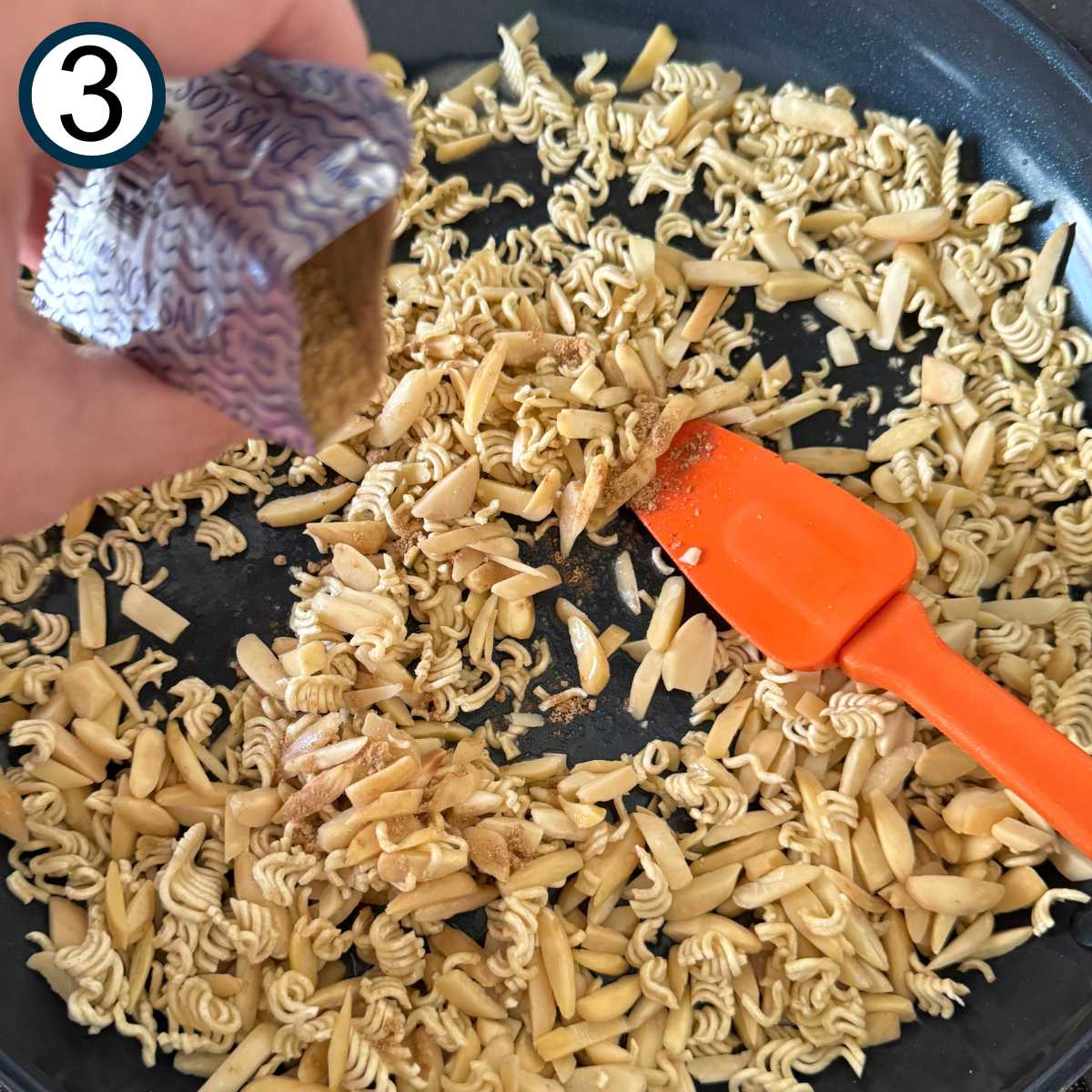 Broken ramen noodles and slivered almonds in a large skillet with a spatula. A seasoning packet is being sprinkled over the top.