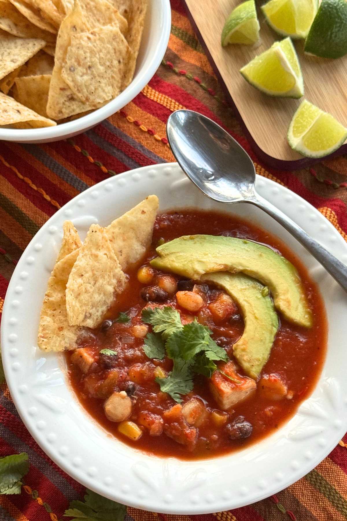 Serving bowl and spoon with chicken tortilla soup showing hominy, black beans, and corn. It's topped with cilantro, 2 slices of avocado, with 3 tortilla chips are tucked on the side. A bowl of tortilla chips and lime wedges are behind it.