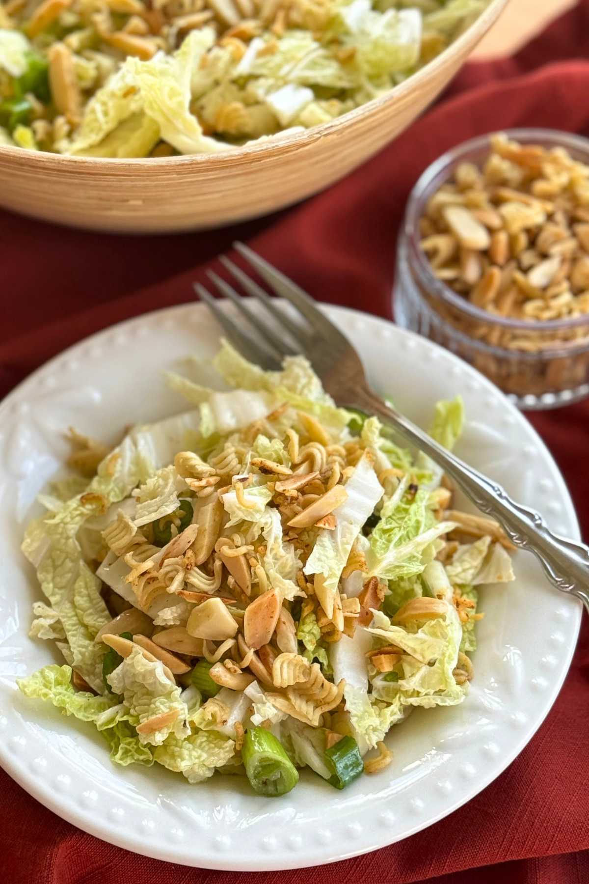 Small bowl of napa cabbage ramen salad with a small glass jar with crispy ramen and nut topping next to it and a large bowl with more salad behind it.